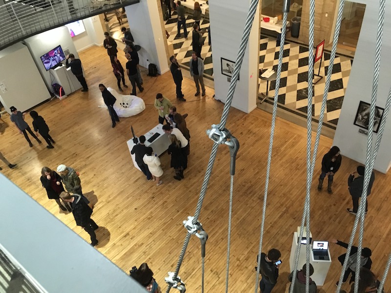A photo taken from overhead showing groups of people clustered around various artworks displayed on small white tables.