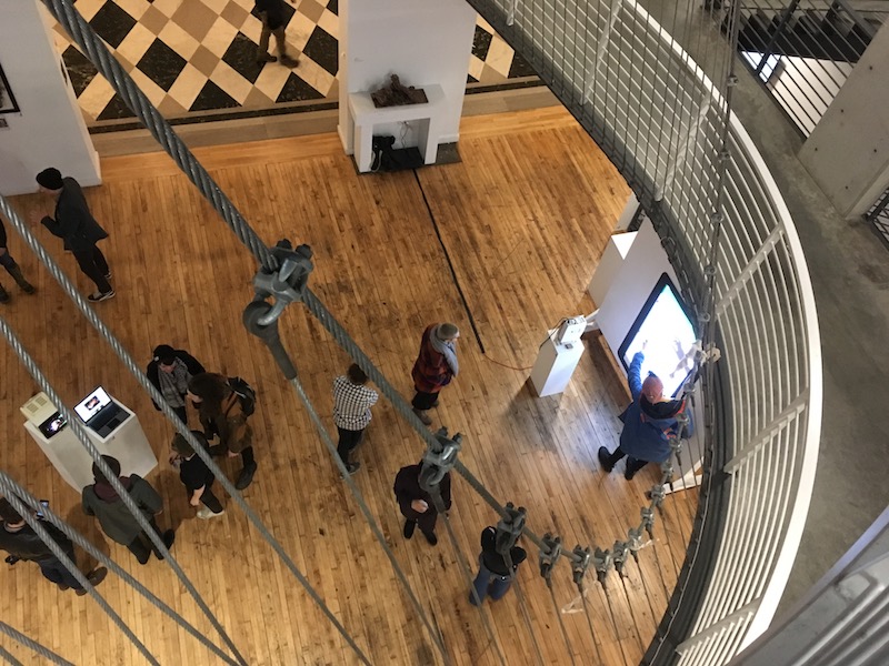 A photo taken from overhead shows groups of people looking at art. One group is clustered around a projector; one person points at the projection.