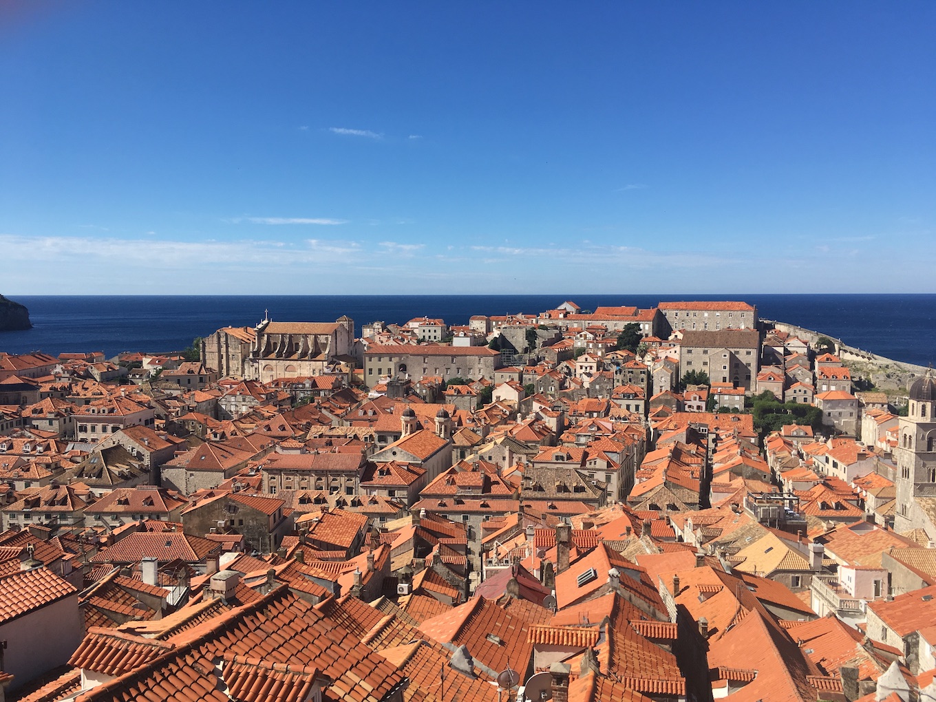 A photo of an old city with narrow streets and orange tiled roofs; the sea is visible beyond.